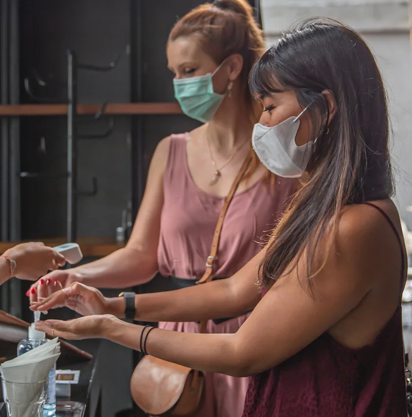 Close-up shot of two multi ethnic female friends following safety protocol of new normal before entering a restaurant. One Caucasian woman getting her temperature check by the receptionist while her Indonesian friends using a hand sanitizer that provided in the entrance counter.