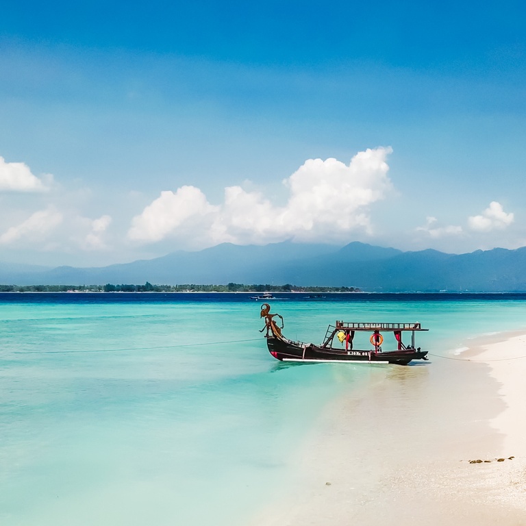 A boat parked next to the shore on Gili Air, Lombok, Indonesia. Beautiful and clear water. In the back visible Mount Rinjani. Some trees on the shore, few clouds on the sky.