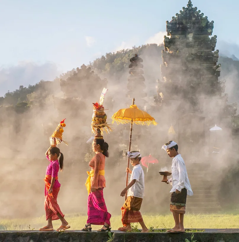 Showing traditional Balinese male and female ceremonial clothing and religious offerings as a mother and children walk to a Hindu temple (pura) in Bali.