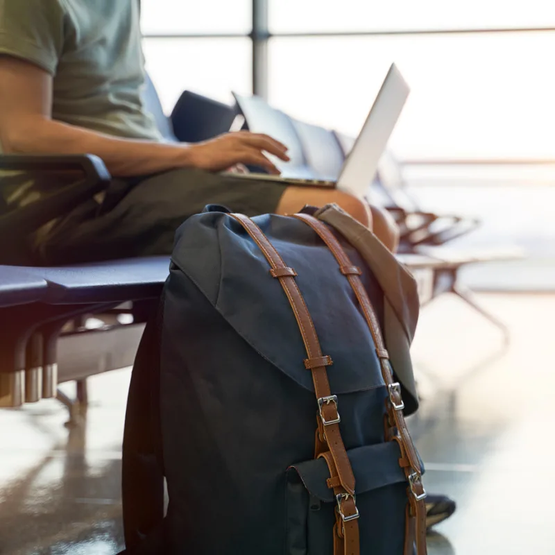 Backpack-Rests-On-The-Floor-Next-To-Man-In-Aiport-Using-Laptop