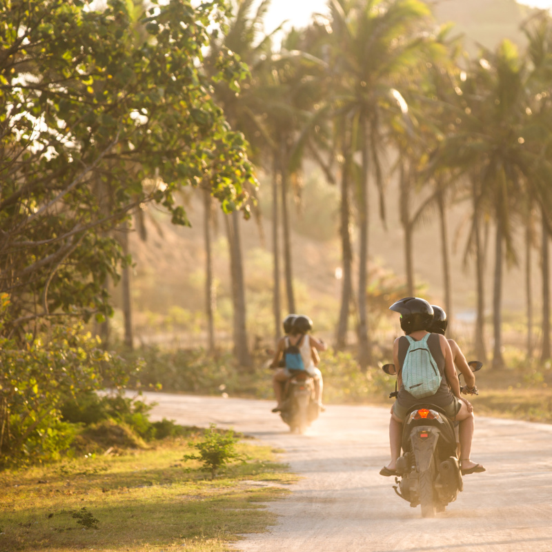 Tourists-In-Bali-Drive-Moped-Scooters-Down-Empty-Road-Lined-With-Palm-Trees