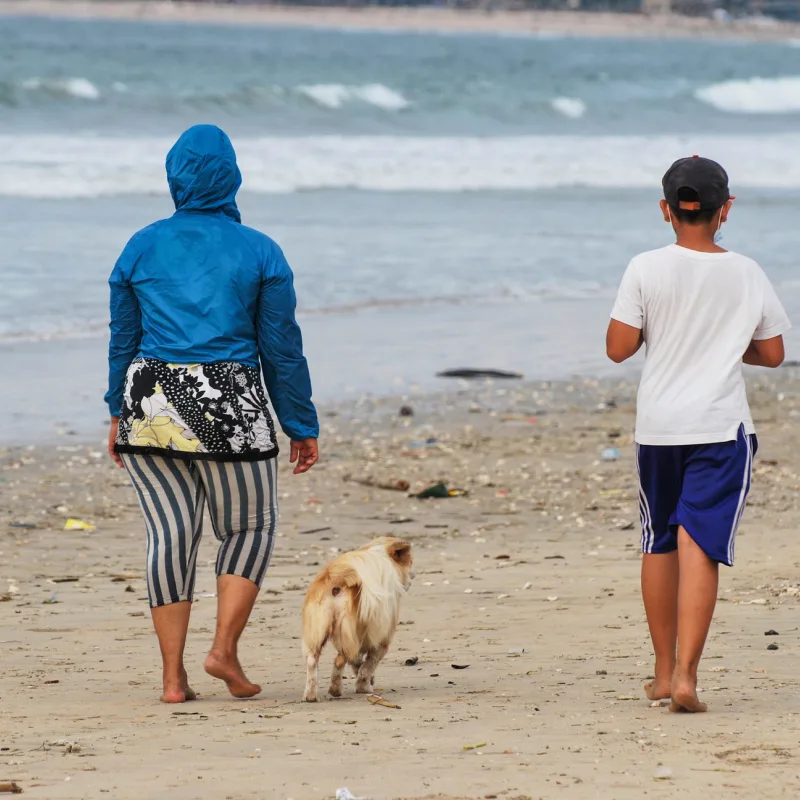 Dua Orang Bali Berjalan di Pantai Bersama Seekor Anjing Putih