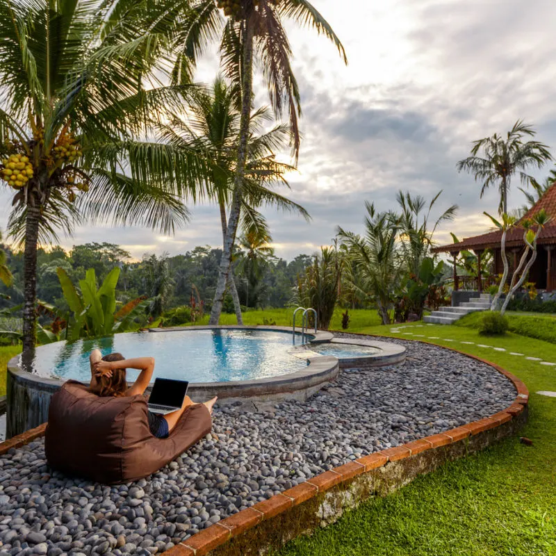 Digital-Nomad-In-Bali-Sits-On-Sun-Lounger-Working-On-Laptop-Surrounded-By-Tropical-Garden-And-Palm-Tree