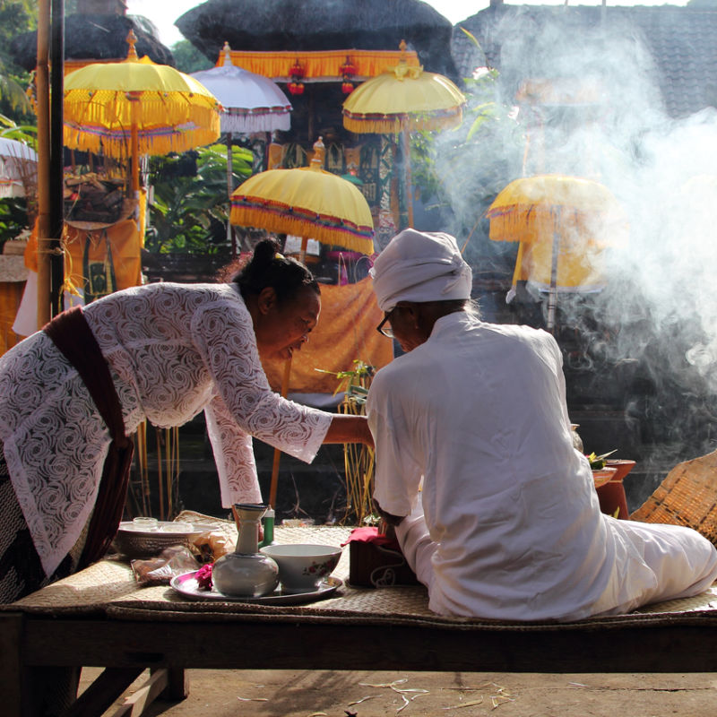 Priest-In-Bali-Does-Cleansing-Ceremony-In-Temple-With-Help-Of-Balinese-Woman-In-Traditional-Dress