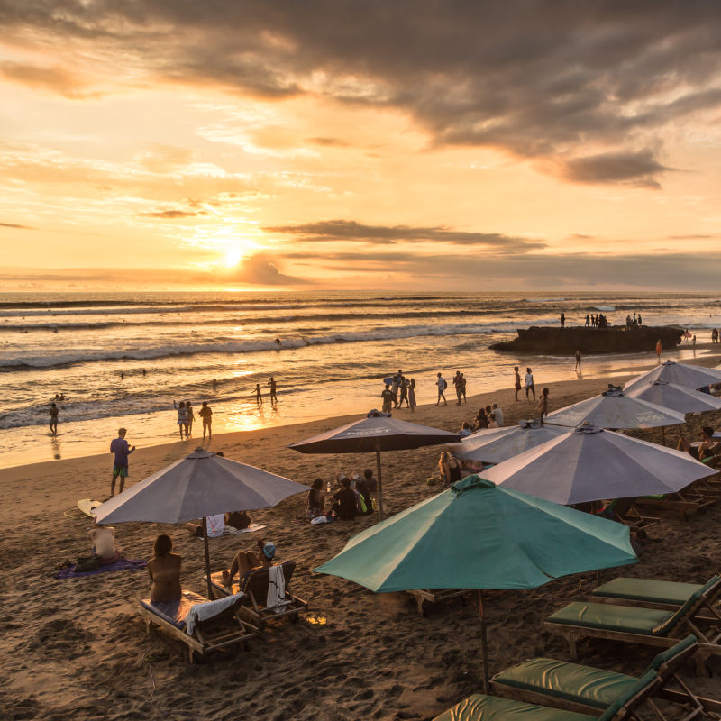 Sunset-on-Seminyak-Beach-In-Bali-Tourists-Play-In-The-Waves-And-Sit-By-Beach-Umbrellas