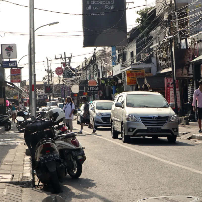 Traffic-On-Street-In-Bali-People-Crossing-The-Road-Past-Cars-and-Moped-Scooters