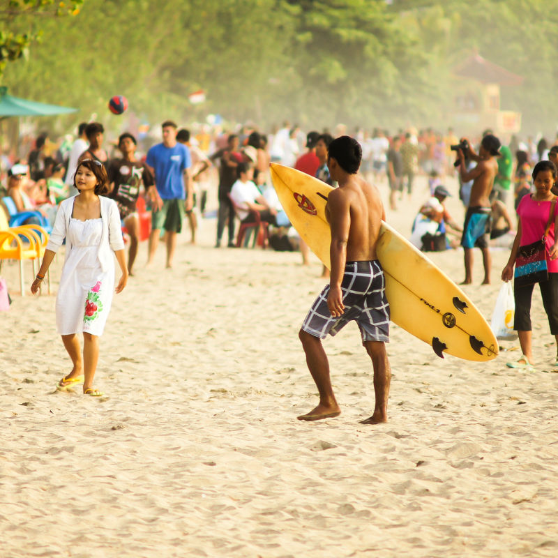 Tourist-Walk-Along-Kuta-Beach-One-Woman-Wears-A-White-Dress-And-A-Man-Carries-A-Yellow-Surfboard