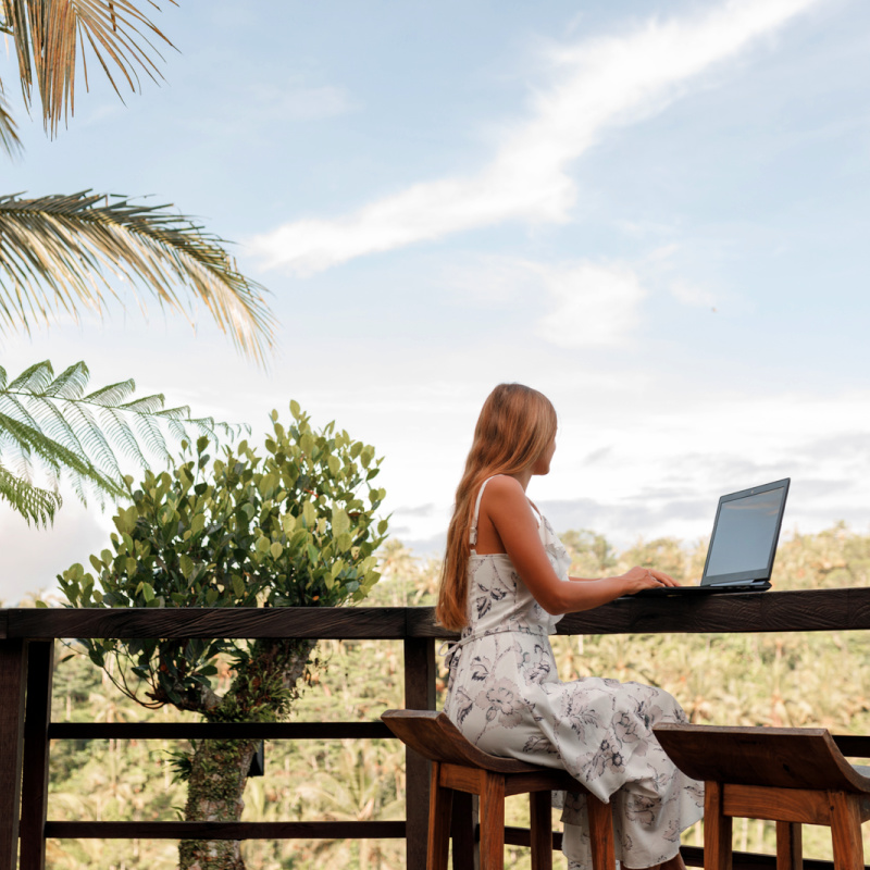 Digital Nomad Sits At Outdoor Work Station In Bali With Laptop Next To Palm Tree