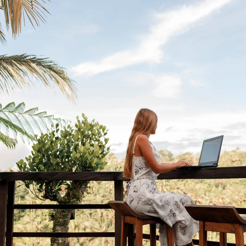 Digital-Nomad-Sits-At-Outdoor-Work-Station-In-Bali-With-Laptop-Next-To-Palm-Tree