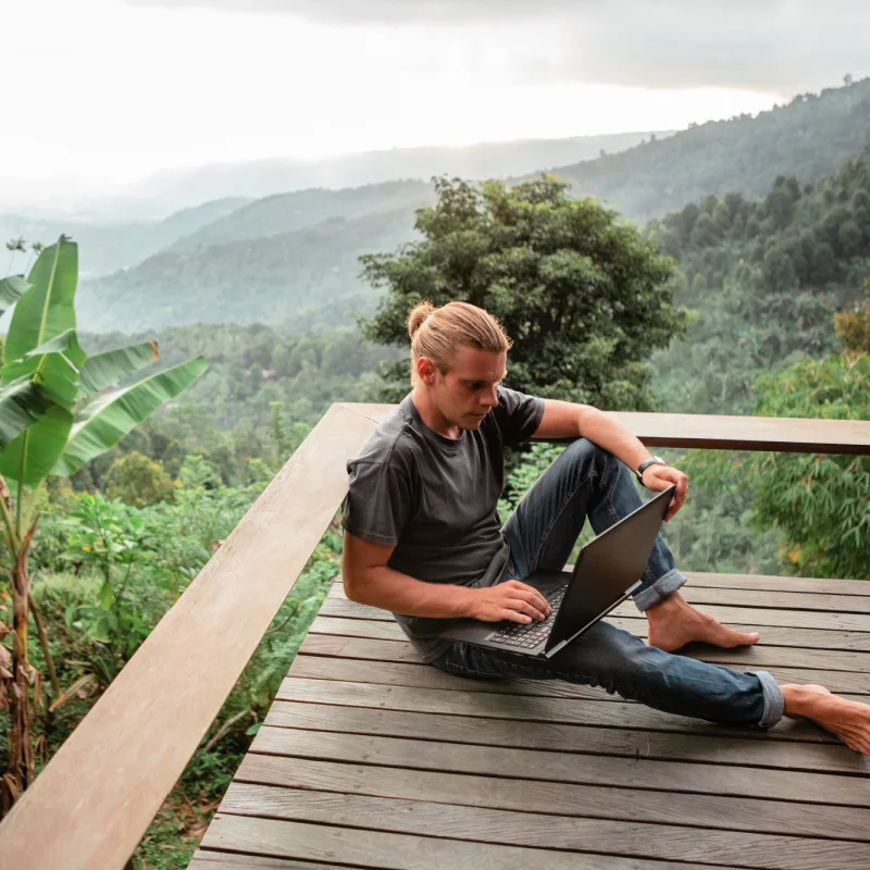 Digital Nomad Sits On Outdoor Wooden Deck In Bali Overlooking Tropical Jungle With Laptop