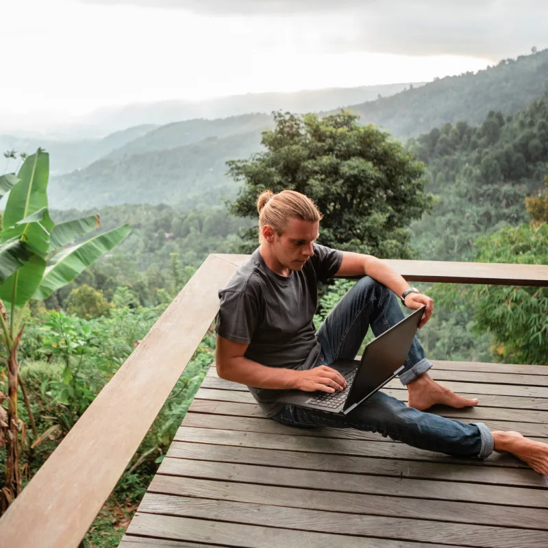 Digital-Nomad-Sits-On-Outdoor-Wooden-Deck-In-Bali-Overlooking-Tropical-Jungle-With-Laptop