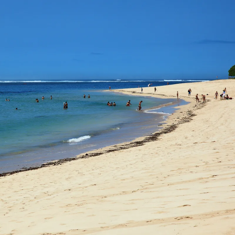 Tourists Relax On The Sand And Shoreline Of Nusa Dua Beach In Bali On Sunny Bright Day