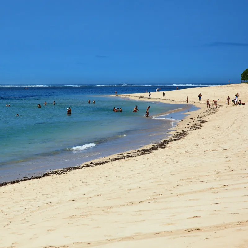 Tourists-Relax-On-The-Sand-And-Shoreline-Of-Nusa-Dua-Beach-In-Bali-On-Sunny-Bright-Day