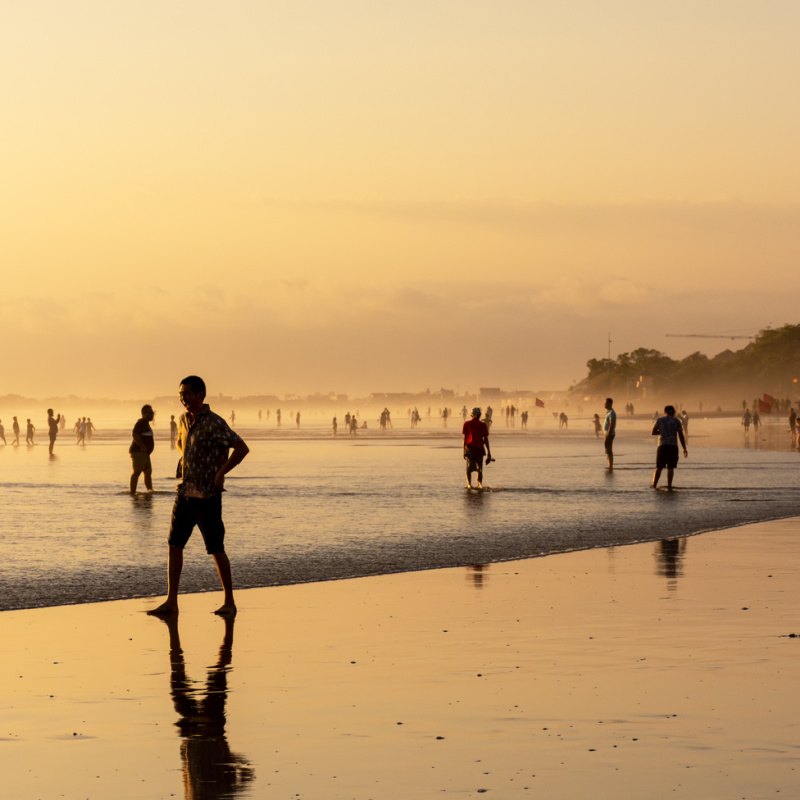 Tourists Walk Down Kuta Beach In Warm Light At Sunset