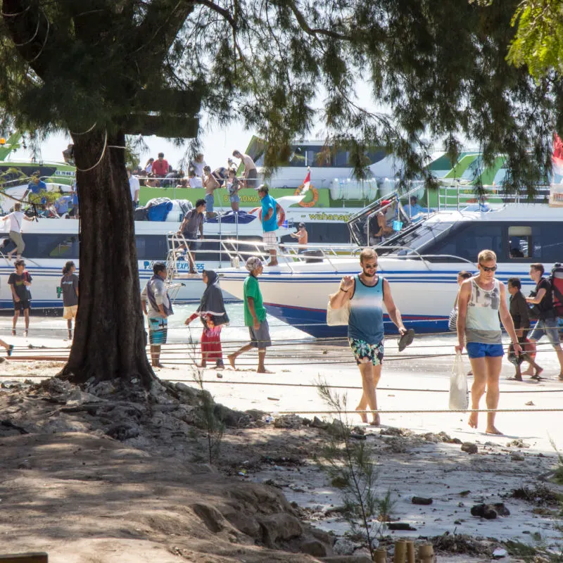 Travlers-On-Gili-T-And-Island-Off-Bali-Walk-Along-Beach-Next-To-Tourist-Fast-Boats