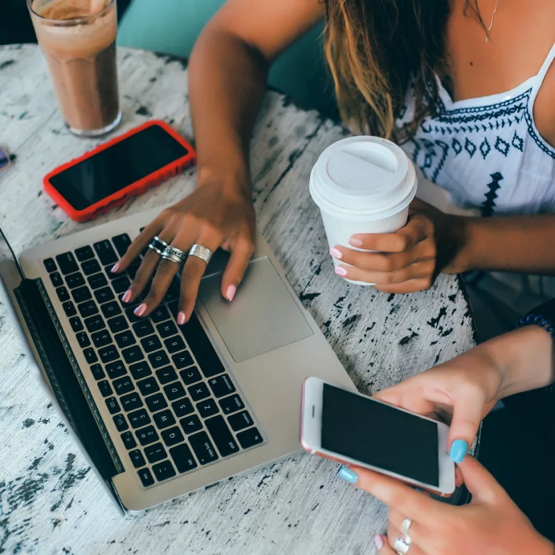 Two Digital Nomads Looking At One Laptop And Their Mobile Cell Phones With A Takeaway Coffee