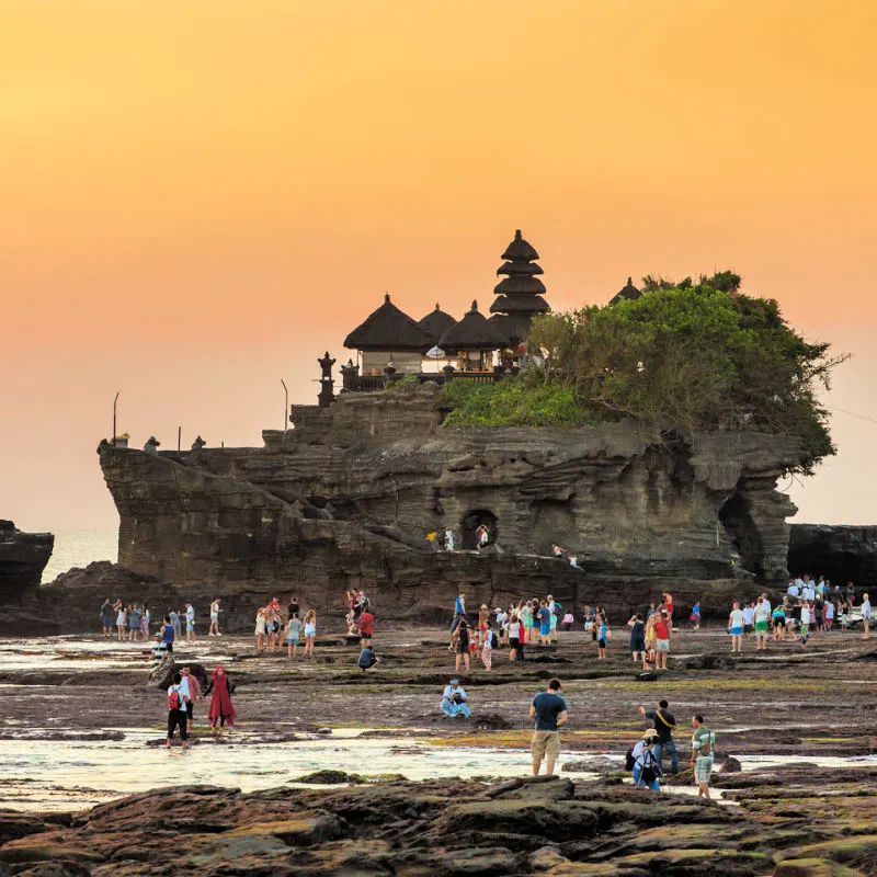 Visitors-Stand-On-Rocky-Shoreline-Looking-Up-At-Tanah-Lot-Temple-In-Bali-Ay-Sunsey