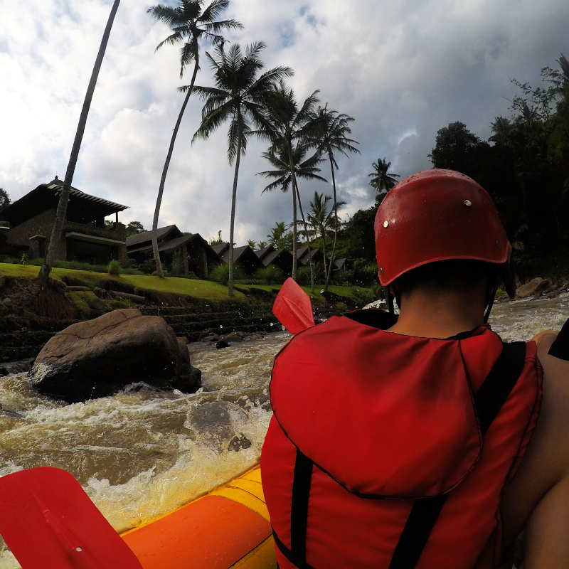 Close-Up-Of-Rafter-From-Behind-Wearing-Red-Life-Jacket-And-Helmet-On-A-Raft-Boat-On-River-In-Bali