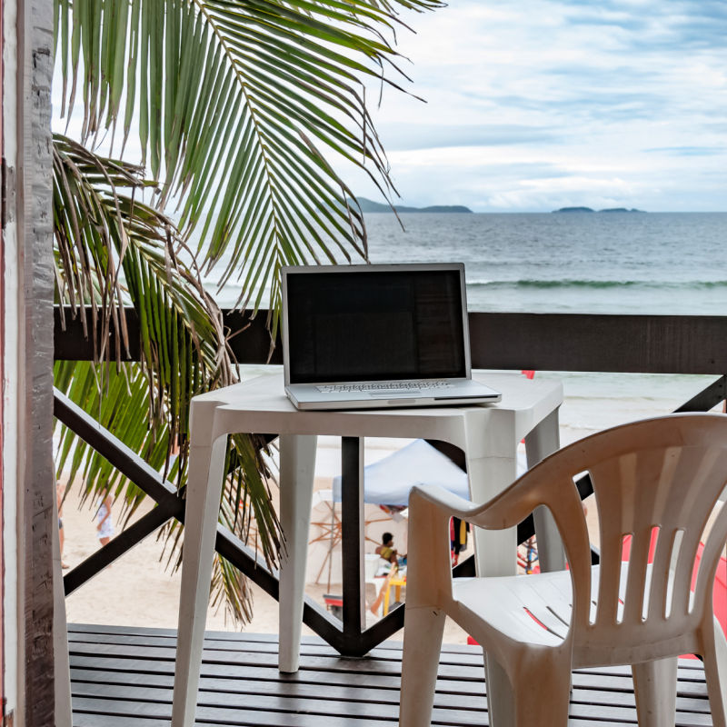 Laptop Sits On A Plastic Table Next To Chair On Wooden Deck With Palm Tree Overlooking Bali Sea View of a digital nomad