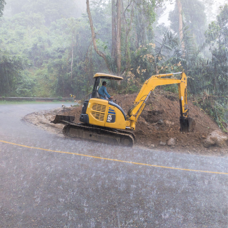 Yellow Excavator Digs At Mud At Side Of Bali Road In The Heavy Rain