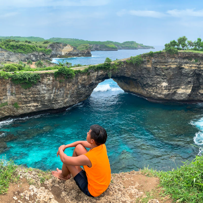 Tourist In Orange Vest Sits At The Edge Of The Cliff At Broken Beach In Bali nusa Penida.
