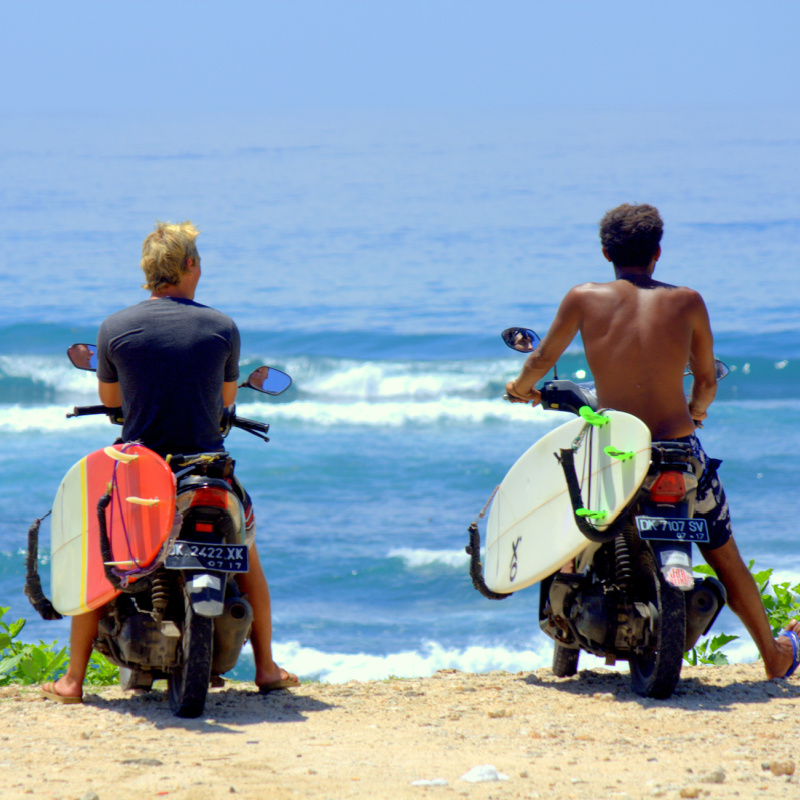 Two Surfers On Mopeds With Surfboards Attached Look Over Cliff In Bali To See Waves