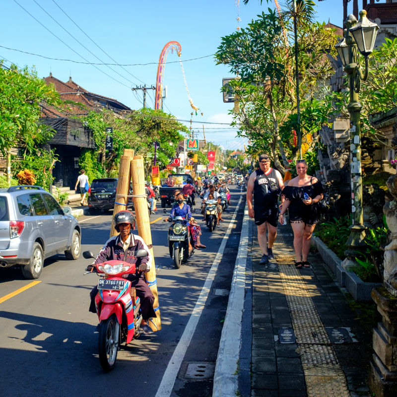 Two-Tourists-Walk-Down-Ubud-High-Street-While-Traffic-Cars-And-Mopeds-Drive-Down-The-Road