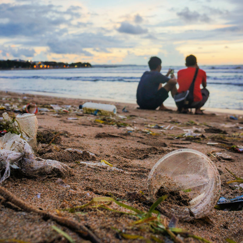 Two Young Men Watch Sunset On Bali Beach That Is Covered in Plastic Waste and Ocean Garbage