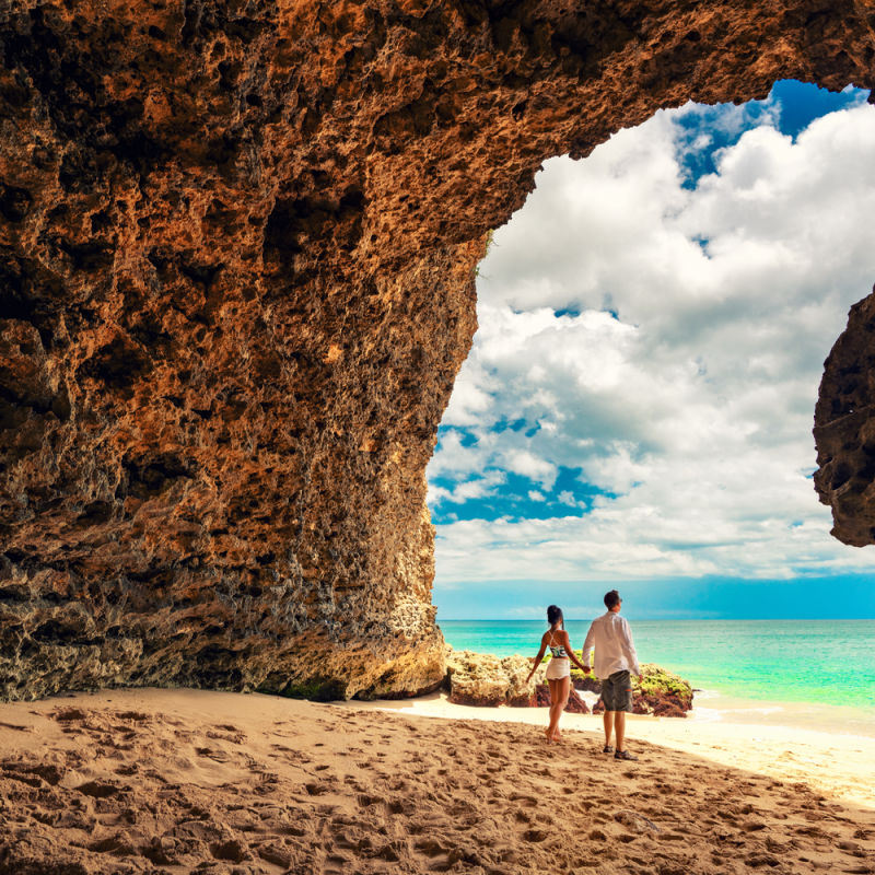 Couple Hold Hands And Look Out At Beach From Cave In Bali