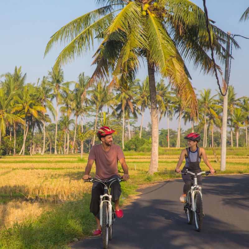 Couple Ride Bikes Along Quiet Bali Road.
