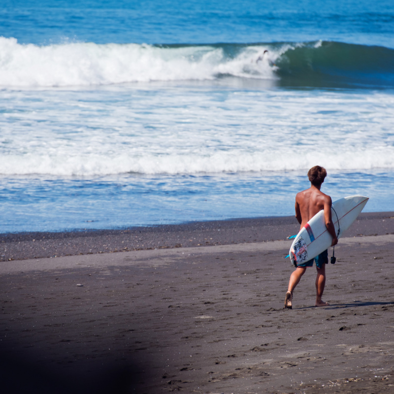 Surfer-Walks-Along-Black-Sand-Bali-Beach-Keramas