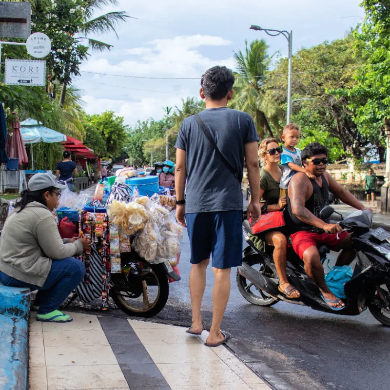 Scene on Bali Street Tourist Walks Family Drives Woman Sells Snacks.jpg