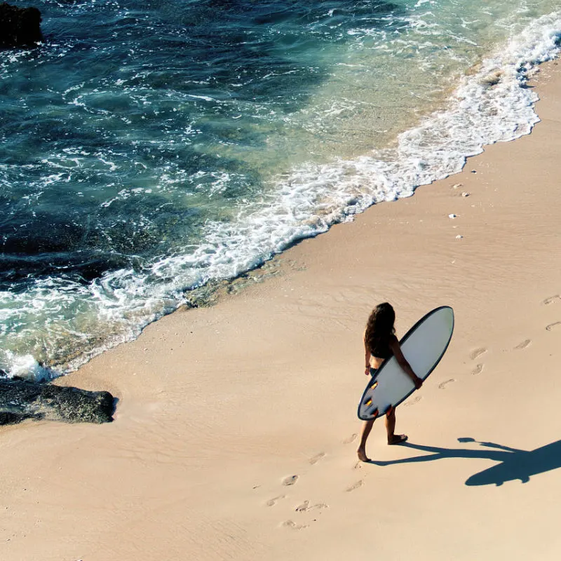 Woman Walks Along Beach With Surfboard